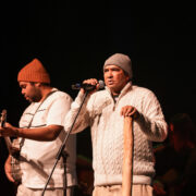 Two men on stage one is playing the guitar and one is speaking into the microphone with his hand on a digeridoo